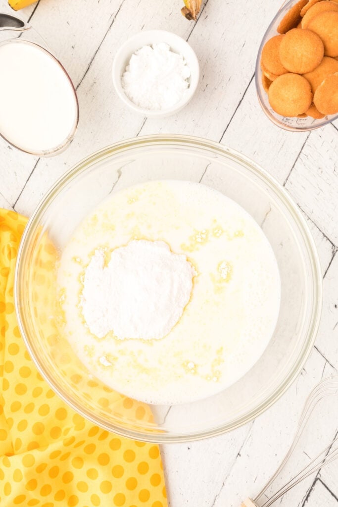 banana pudding mix added to milk in a glass mixing bowl with measuring cups and cookies nearby