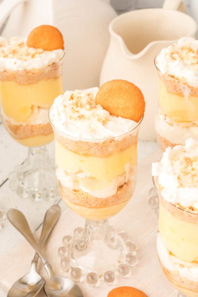 banana pudding parfait layered in glass cups with whipped topping and vanilla wafer on top with spoons and pitcher in the background