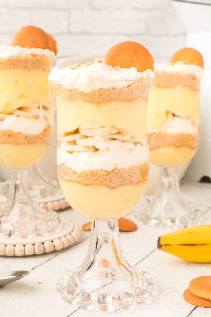 banana pudding parfait layered with sliced bananas, whipped topping, and cookie crumbs in a glass cup with whole bananas and vanilla wafers nearby