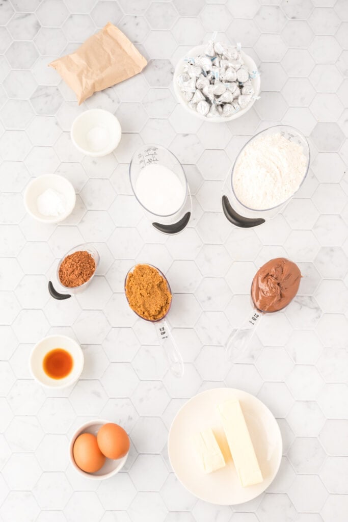 Ingredients for Chocolate Blossoms arranged on a white hexagon tile countertop including flour, sugars, Nutella, cocoa powder, eggs, butter, vanilla, chocolate pudding mix, and Hershey's Kisses