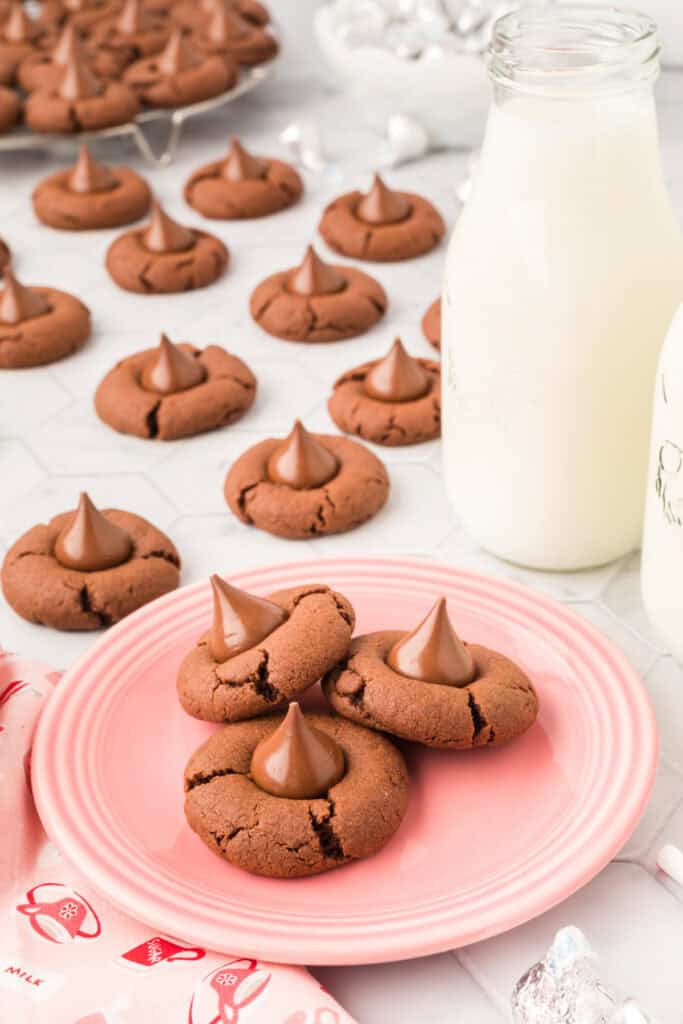 Three Chocolate Blossoms cookies topped with Hershey's Kisses on a pink plate with bottles of milk in the background