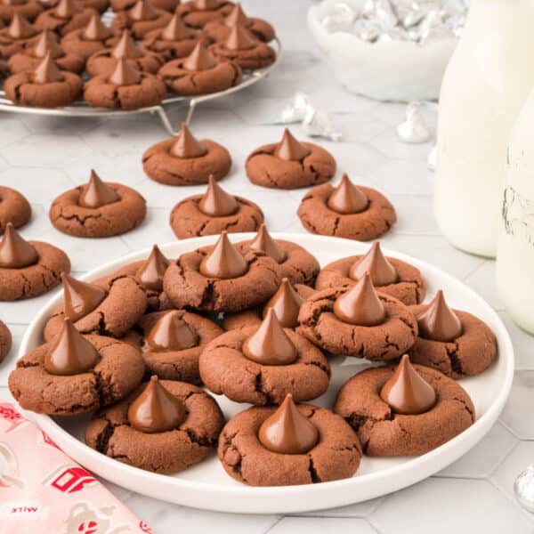 Chocolate Blossoms cookies topped with Hershey's Kisses arranged on a white plate with milk and additional cookies in the background