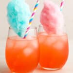 Two vibrant cotton candy margaritas in stemless glasses, one topped with blue cotton candy and the other with pink, set on a light neutral background with pastel striped straws.