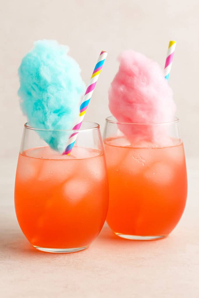 Two vibrant cotton candy margaritas in stemless glasses, one topped with blue cotton candy and the other with pink, set on a light neutral background with pastel striped straws.