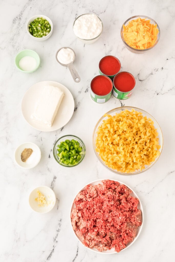 ingredients for beefy sour cream noodle bake arranged on a marble surface including ground beef, egg noodles, tomato sauce, cheeses, and chopped vegetables