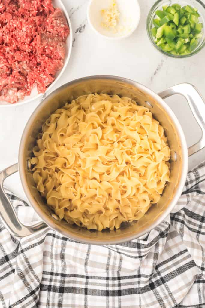 cooked egg noodles in a large pot with raw ground beef and chopped green pepper nearby