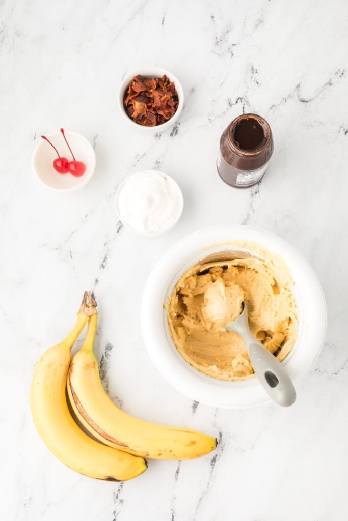 Ingredients for making an Elvis Banana Split, including bananas, homemade peanut butter ice cream, hot fudge sauce, bacon, whipped cream, and maraschino cherries.