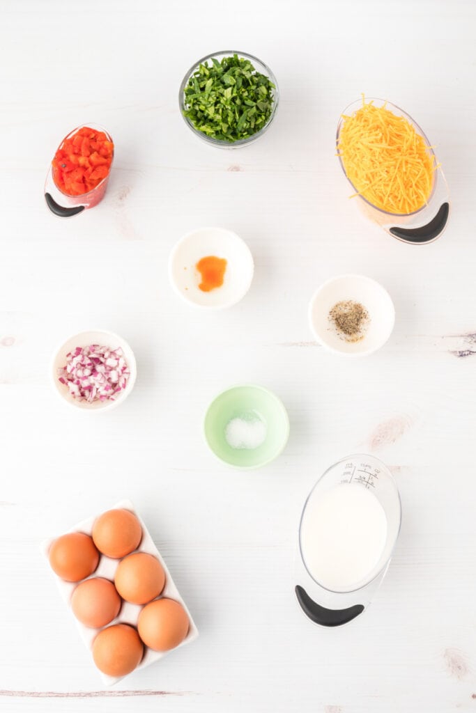 Ingredients for Breakfast Egg Muffins including eggs, milk, spinach, roasted red peppers, red onion, and shredded cheese on a white surface