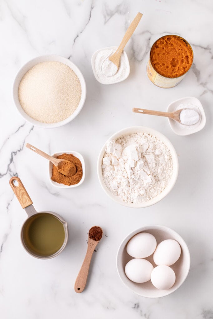 Overhead view of measured ingredients for pumpkin bars including flour, sugar, pumpkin puree, eggs, oil, and spices on a marble countertop.