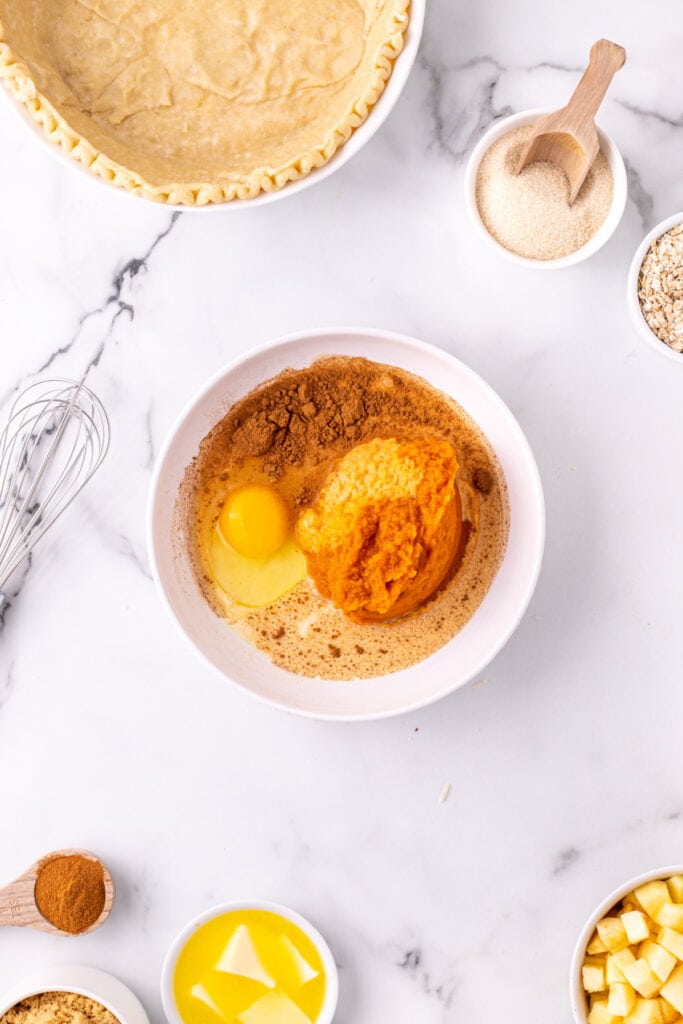 Bowl with pumpkin pie filling, egg, spices, and evaporated milk being prepped next to an unbaked pie crust.
