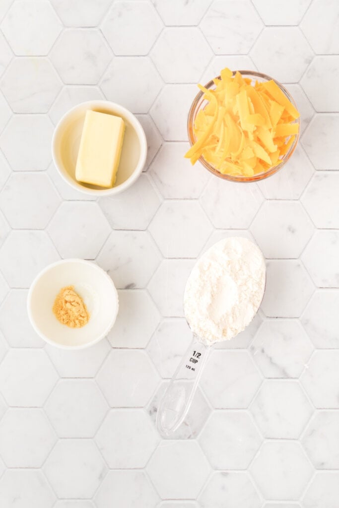Overhead view of ingredients for homemade cheese bites including Cheddar cheese, butter, flour, and ground mustard on a white tile surface