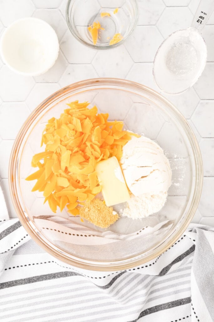 Ingredients for easy cheese bites recipe in a glass mixing bowl with shredded Cheddar cheese, flour, butter, and ground mustard