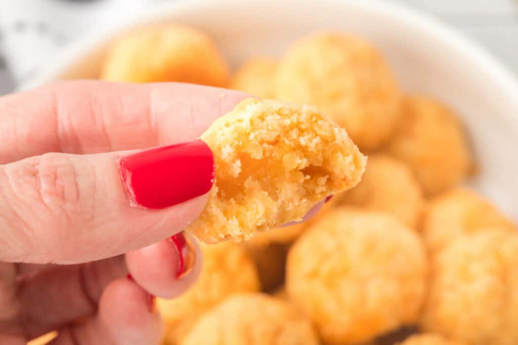 Hand holding a homemade cheese bite with a soft cheesy center and crumbly texture over a bowl of baked cheese bites