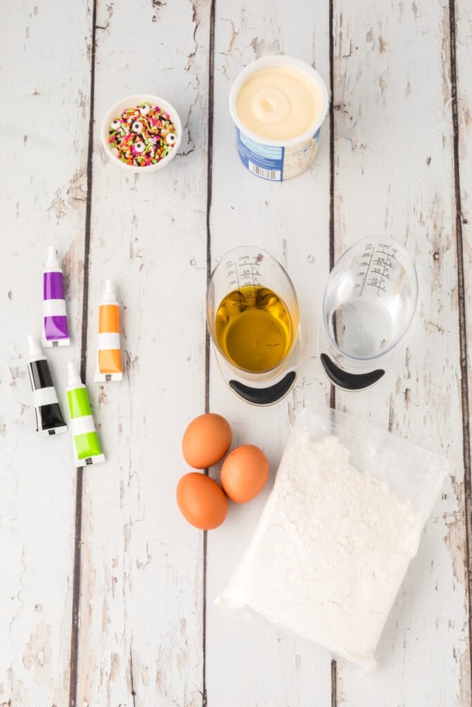 Overhead view of Halloween Bundt Cake ingredients including cake mix, eggs, oil, water, frosting, sprinkles, and food coloring.