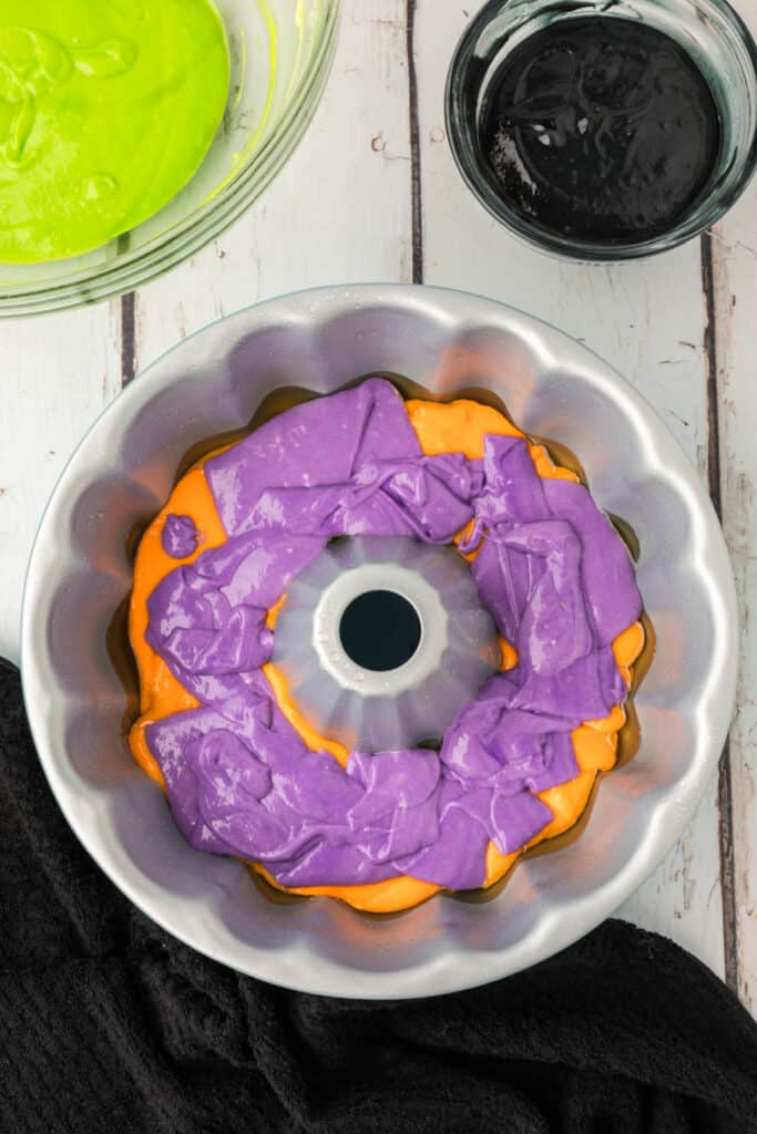 Purple cake batter layered on top of orange batter in a bundt pan for Halloween Bundt Cake.