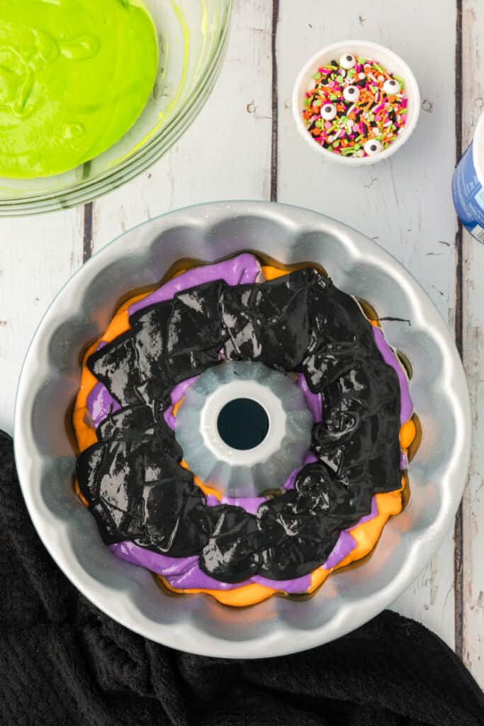 Bundt pan with layered orange, purple, and black cake batter, with a bowl of green batter and Halloween sprinkles nearby.