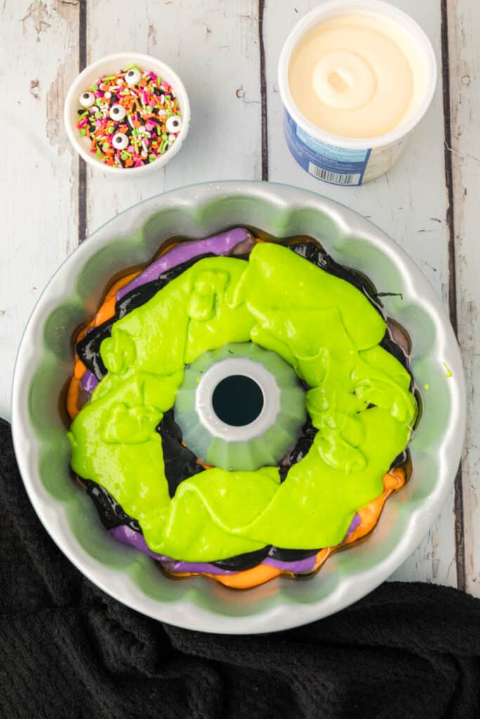Bundt pan filled with green cake batter on top of purple, black, and orange layers for Halloween Bundt Cake.