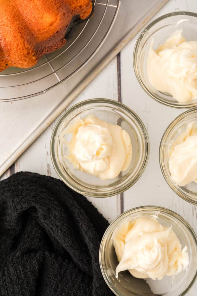 Bowls of white frosting divided for coloring beside cooled Halloween Bundt Cake.