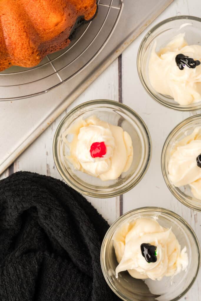 Small bowls of frosting with drops of food coloring ready to be mixed for cake drizzle.
