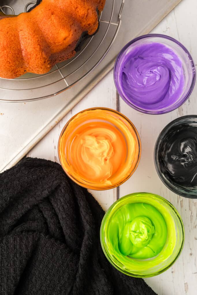 Bowls of tinted frosting in orange, purple, green, and black next to cooled bundt cake.