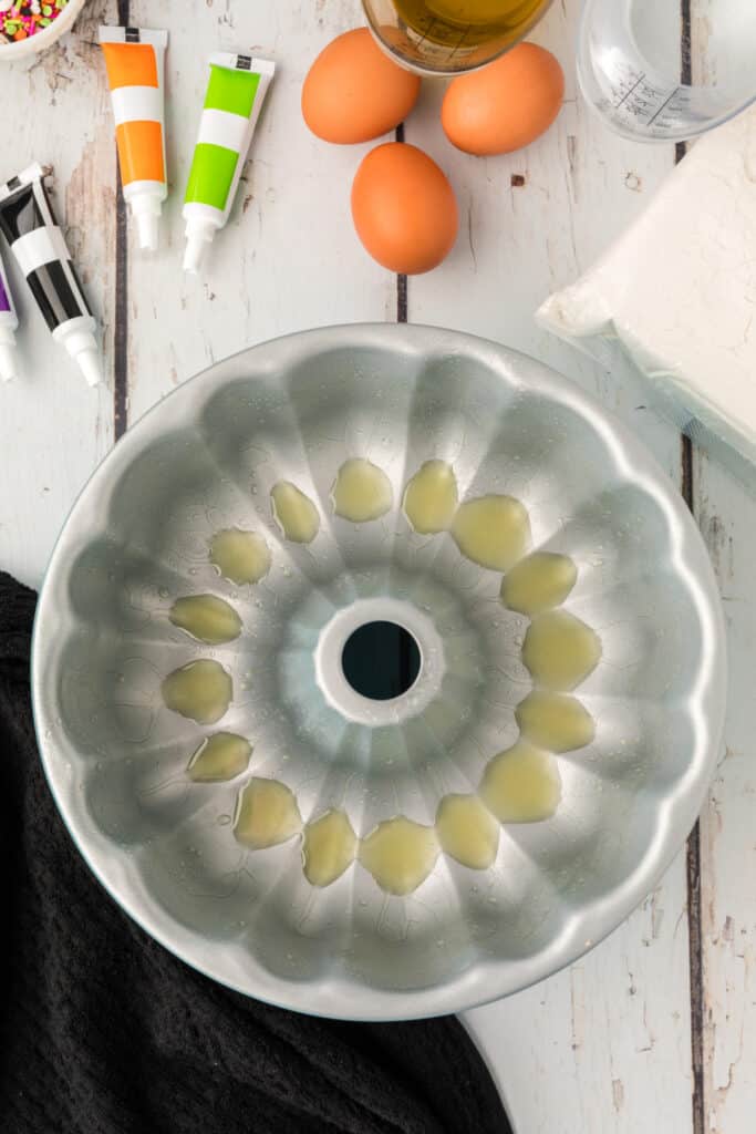 Greased bundt pan surrounded by Halloween Bundt Cake ingredients like eggs, oil, flour mix, and colorful food coloring tubes.
