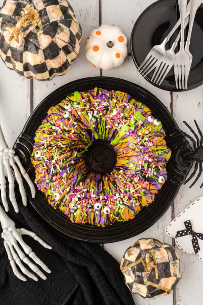Decorated Halloween Bundt Cake on a black platter surrounded by pumpkins, forks, and skeleton hand props