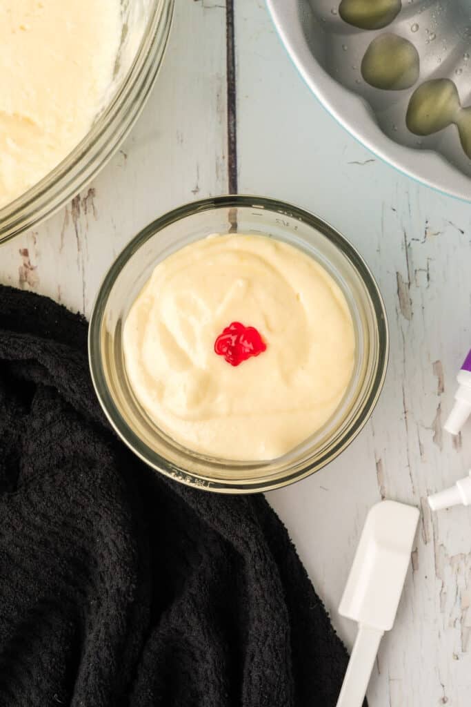 Small bowl of white cake batter with a drop of red food coloring added to create orange batter.