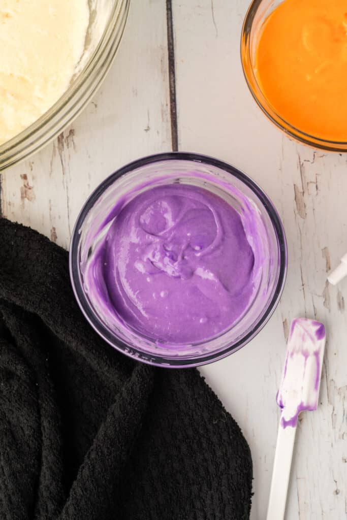Bowl of purple cake batter with spatula streaked in matching color beside it on a black kitchen towel.