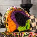 Square image of sliced Halloween Bundt Cake with orange, purple, green, and black layers topped with sprinkles.