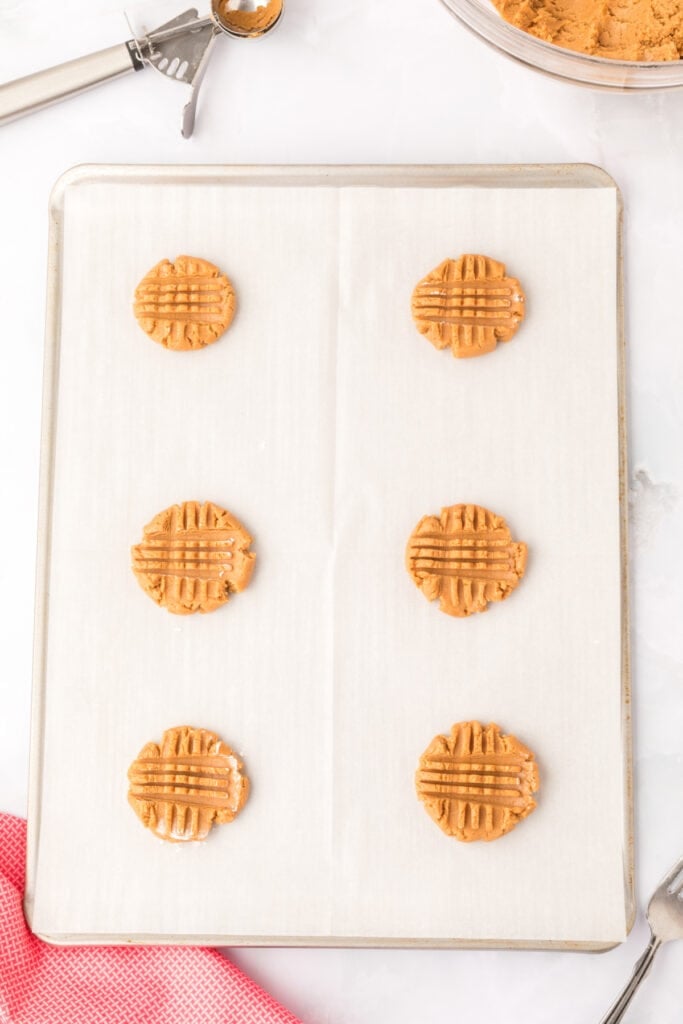 six unbaked biscoff cookie dough balls flattened with fork marks on parchment-lined baking sheet with metal cookie scoop and bowl of dough partially visible