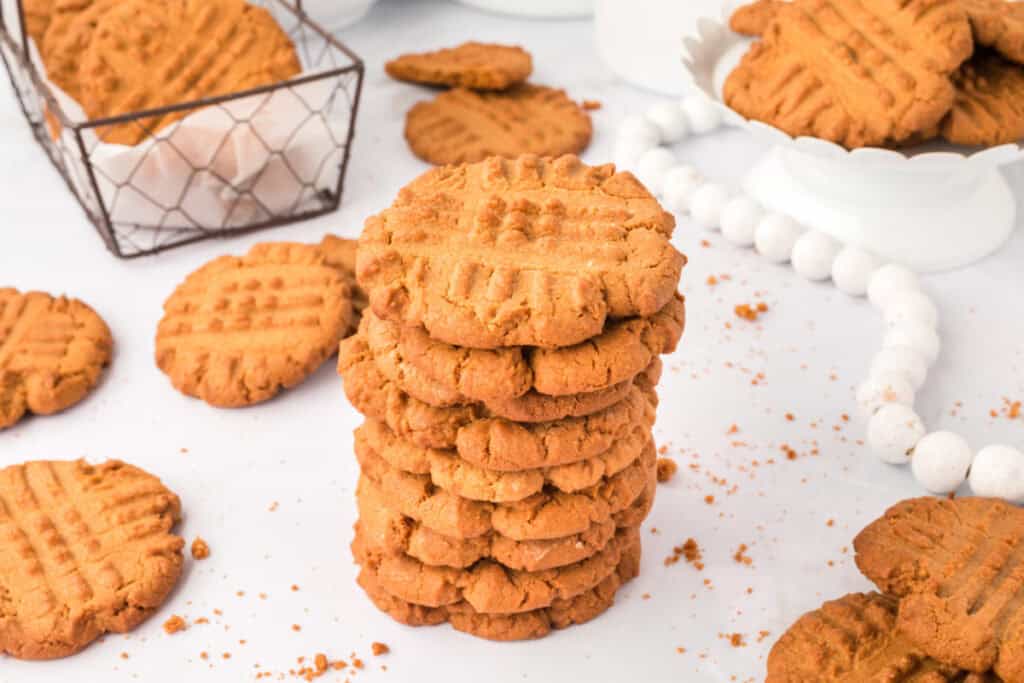 biscoff butter cookies piled on white surface with crumbs, a basket of cookies, a white plate of cookies, and a jar of lotus biscoff cookie butter