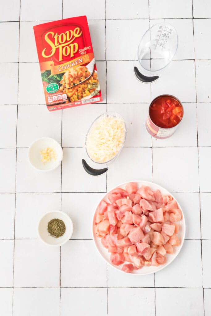 Ingredients for Chicken Bruschetta Bake arranged on a white tiled surface, including diced chicken, mozzarella cheese, diced tomatoes, stuffing mix, garlic, basil, and water.