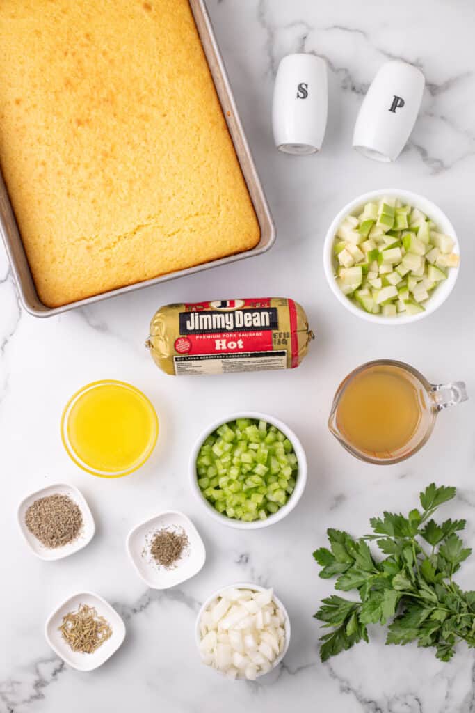 Overhead view of baked cornbread in a pan with bowls of chopped apples, celery, onions, sausage, herbs, melted butter, and broth arranged on a marble surface.