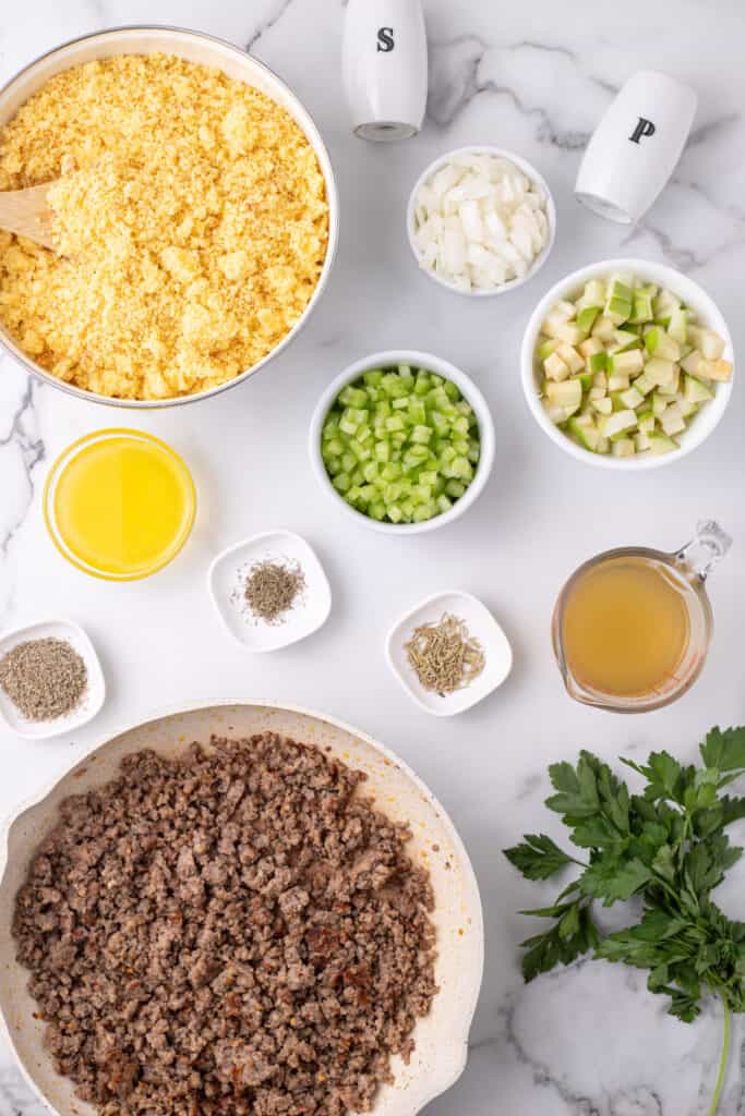 Top down image of crumbled cornbread in a large bowl surrounded by cooked sausage, chopped veggies, herbs, melted butter, and broth.