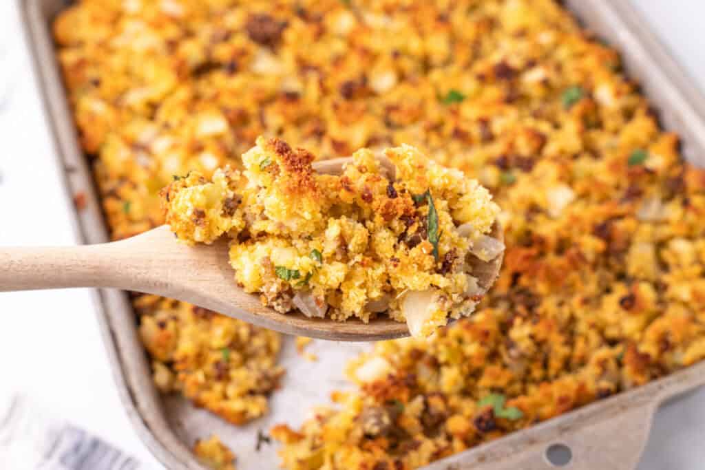 Close up of a wooden spoon lifting a serving of golden brown Southern cornbread dressing from the baking dish, showing the crumbly texture and sausage pieces.