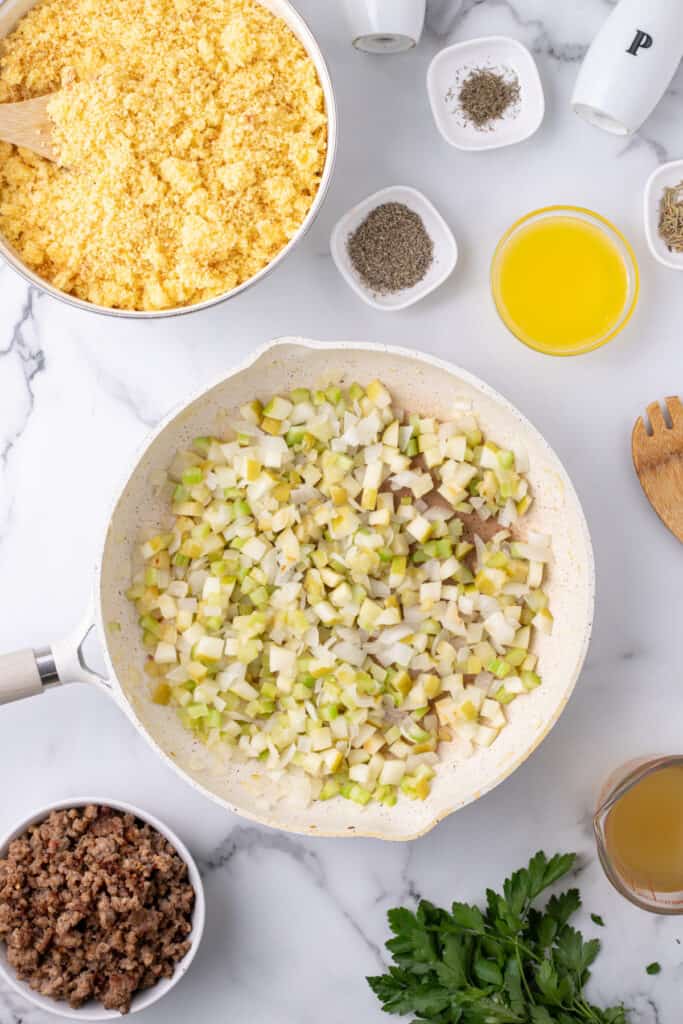 Sautéed onions, celery, and apples in a skillet next to ingredients for cornbread dressing including crumbled cornbread and spices.