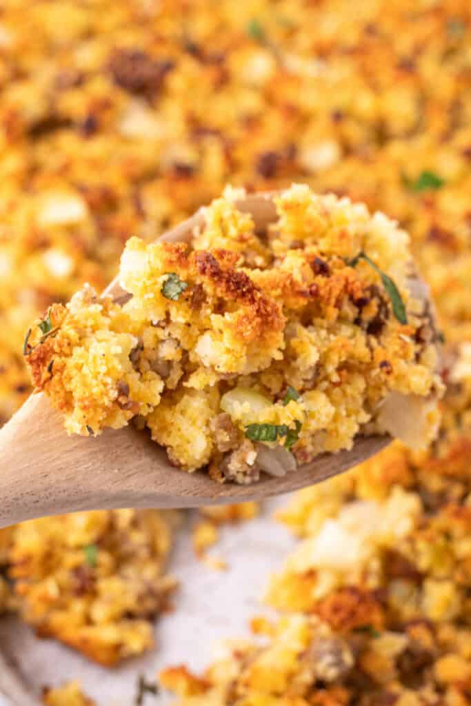 Close up of a wooden spoon holding a scoop of golden brown Southern cornbread dressing with browned edges, sausage, onions, and fresh herbs.