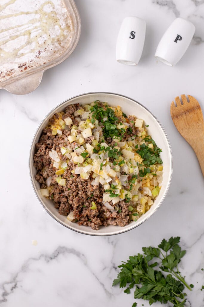 Large mixing bowl filled with crumbled cornbread topped with cooked sausage, sautéed veggies, and chopped parsley before mixing.