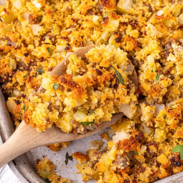 Close up square photo of Southern cornbread dressing with a wooden spoon scooping up a portion, showing crispy browned bits and soft cornbread pieces.