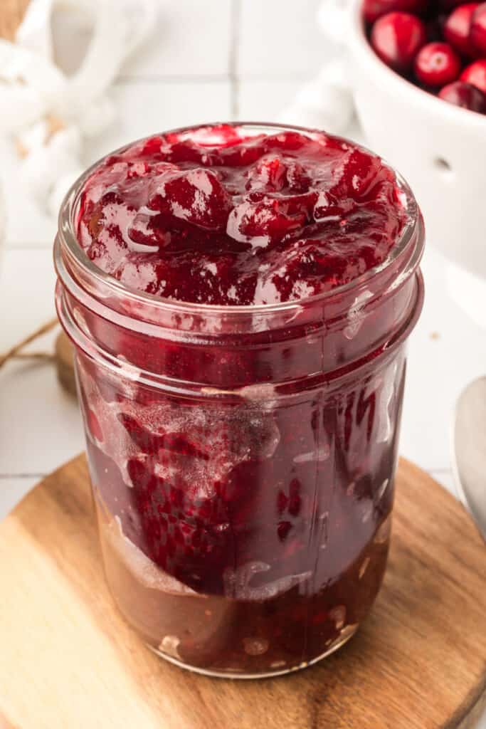Jar of homemade cranberry jam on a wooden board with fresh cranberries and biscuits nearby.