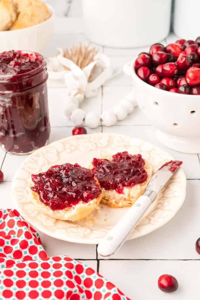 Plate with a biscuit sliced open and spread with cranberry jam, surrounded by fresh cranberries and a jar of jam.