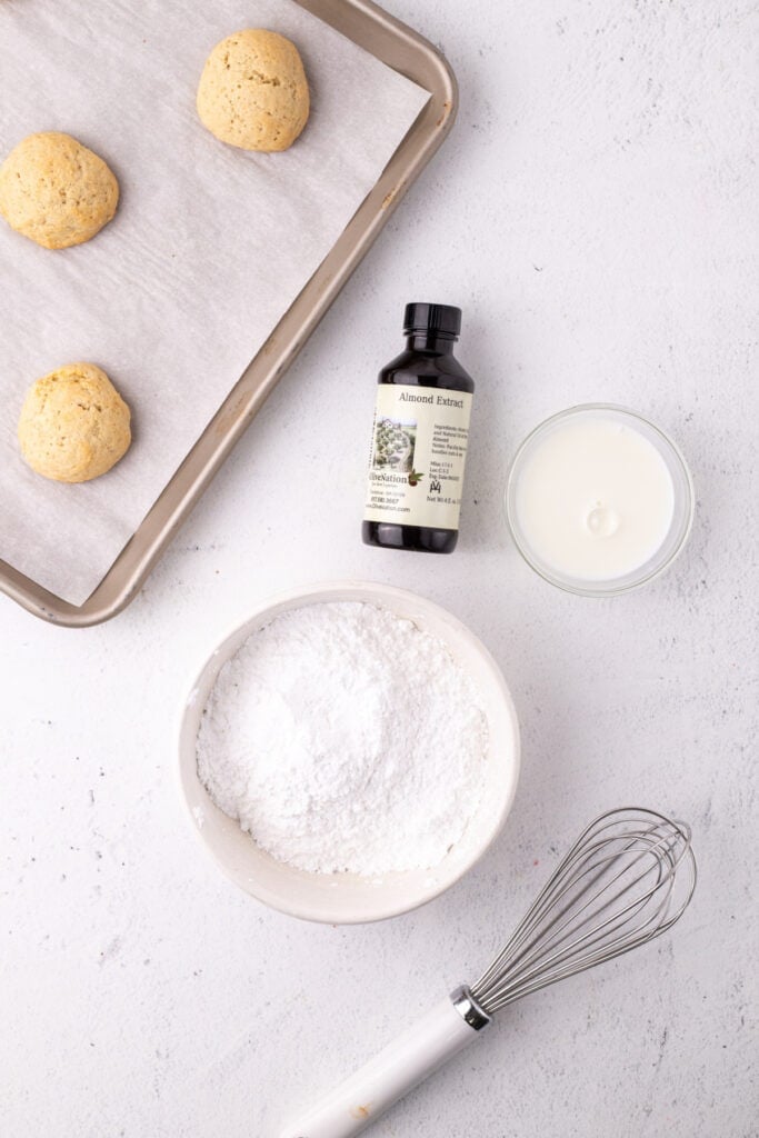 Almond glaze ingredients next to cooled ricotta cookies on baking tray
