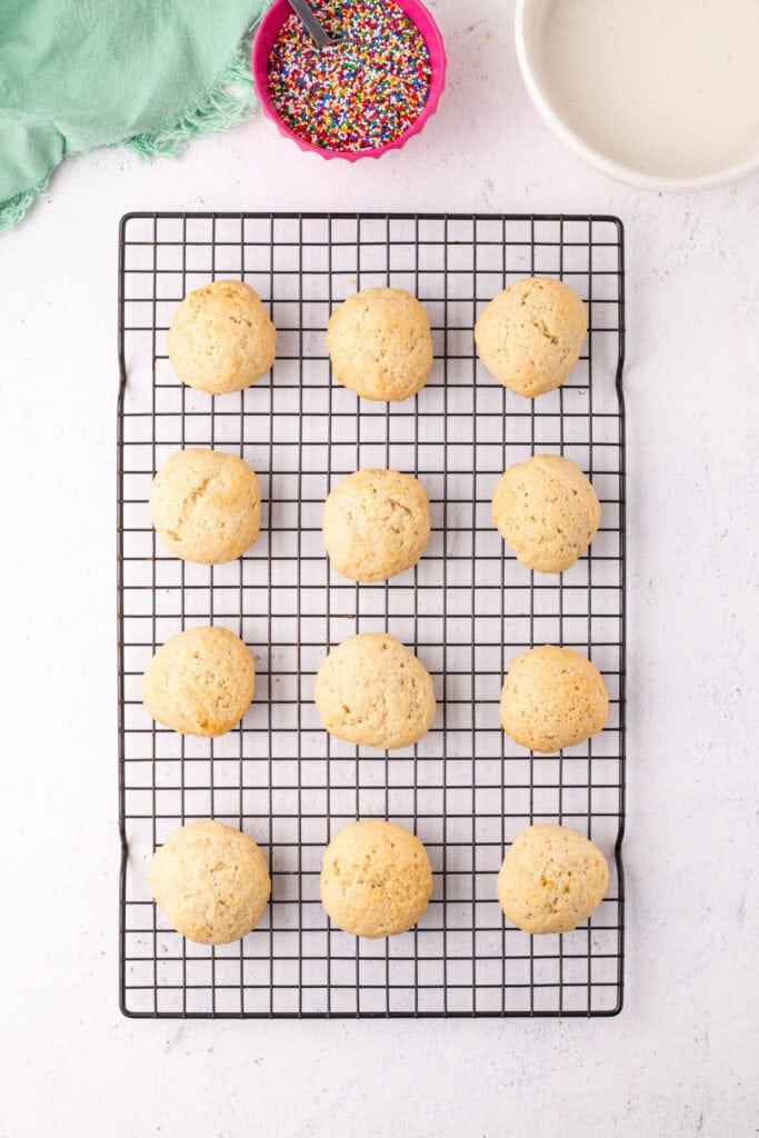 Baked ricotta cookies cooling on a wire rack with glaze and sprinkles nearby