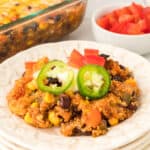 square image of black bean corn enchilada quinoa casserole served on a plate with jalapeno slices, sour cream, and diced tomatoes