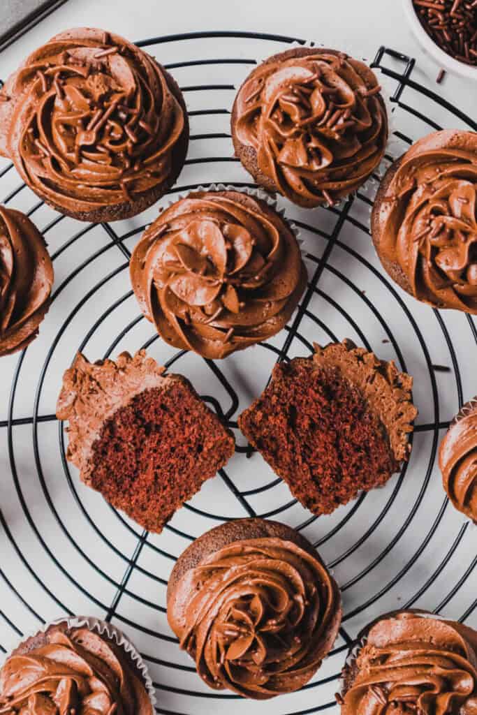 Frosted chocolate cupcakes with chocolate sprinkles on a cooling rack, including a cupcake cut in half