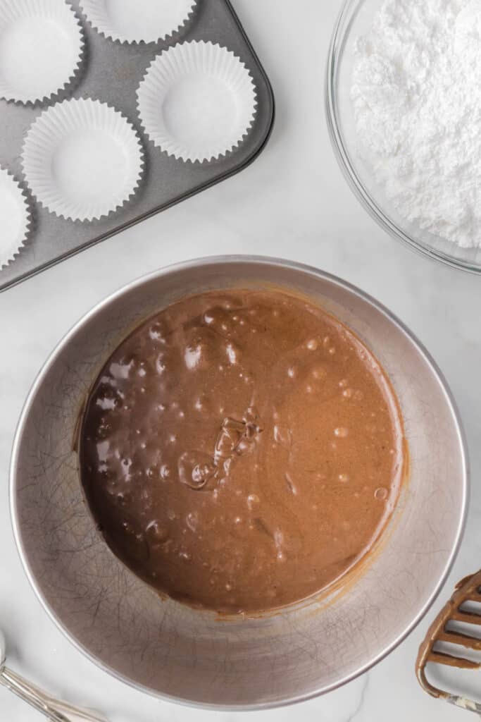 Thin chocolate cupcake batter in a mixing bowl beside cupcake liners and frosting ingredients