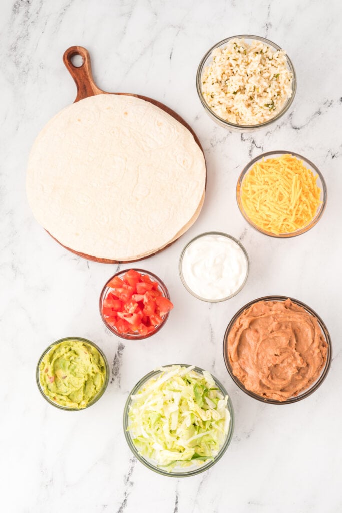 flour tortillas on a wooden board with bowls of refried beans, shredded cheddar cheese, sour cream, guacamole, chopped tomatoes, lettuce, and cilantro lime rice