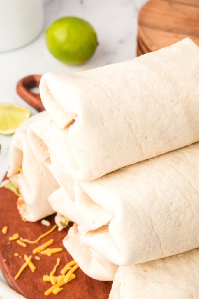 stack of rolled burritos on a wooden board with shredded cheese scattered nearby