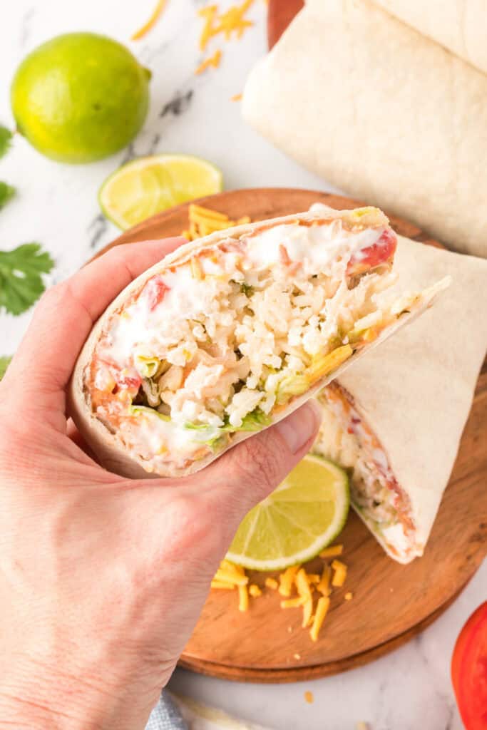 hand holding a cut burrito showing layers of rice, beans, sour cream, cheese, and vegetables with lime slices on a wooden board