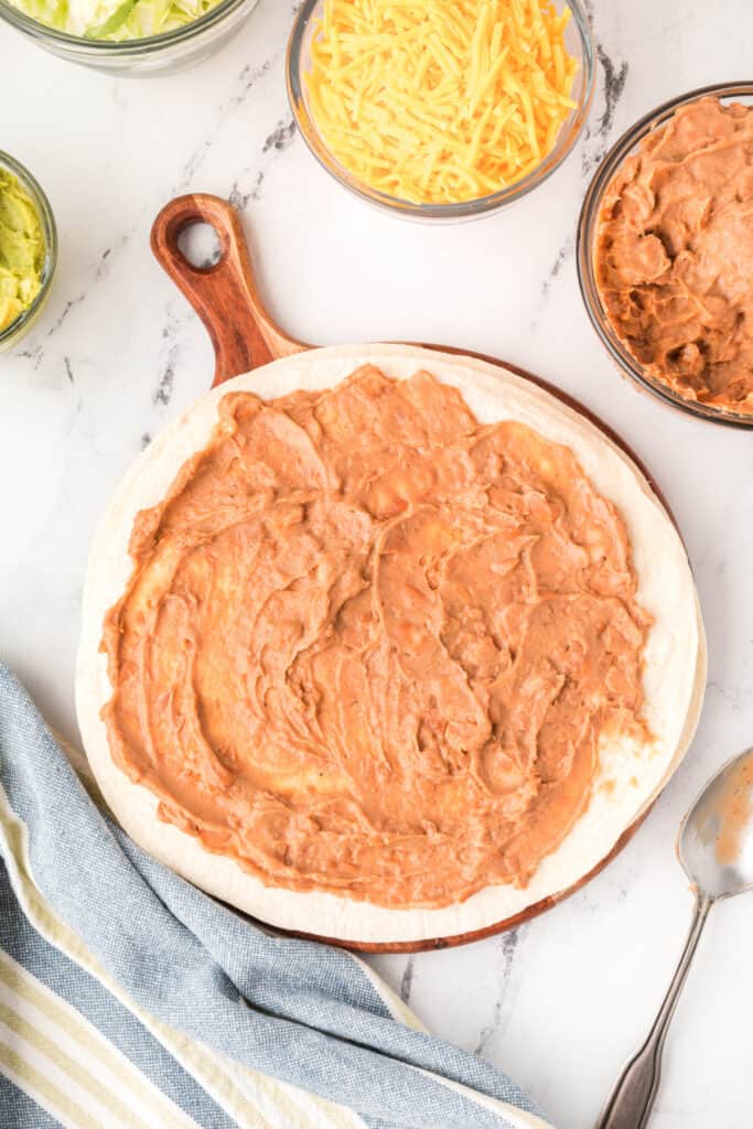 flour tortilla on a wooden board spread with refried beans with bowls of cheese and beans nearby
